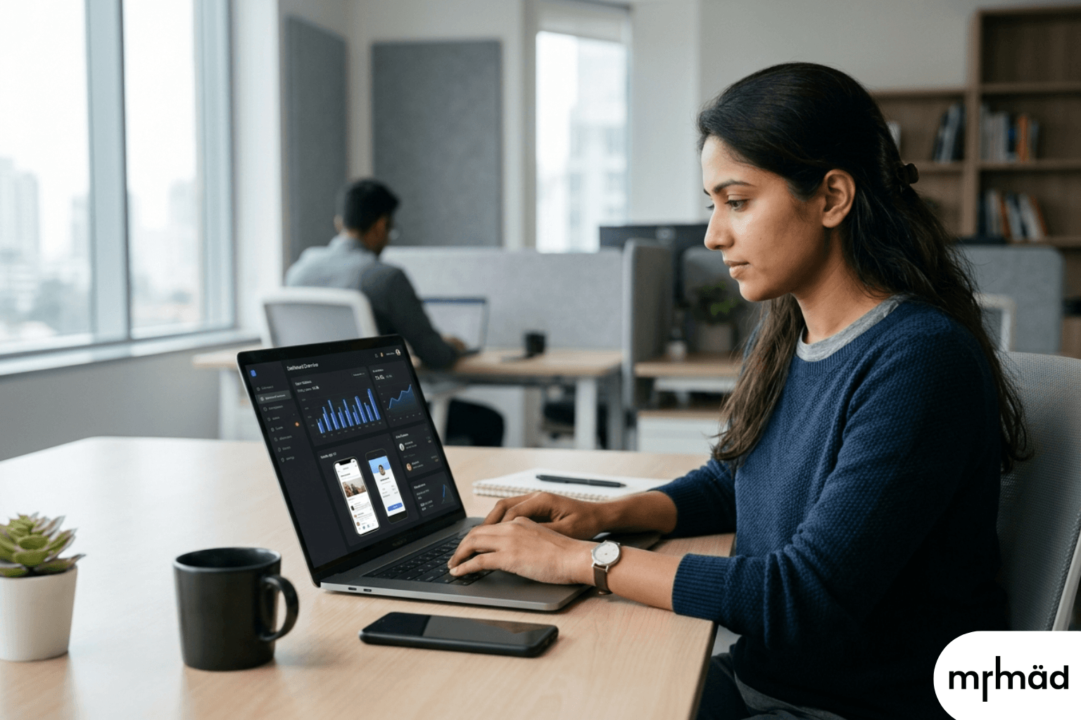 A focused Indian female software developer in a modern office, working on a dark-mode SaaS analytics dashboard and mobile app on her laptop.